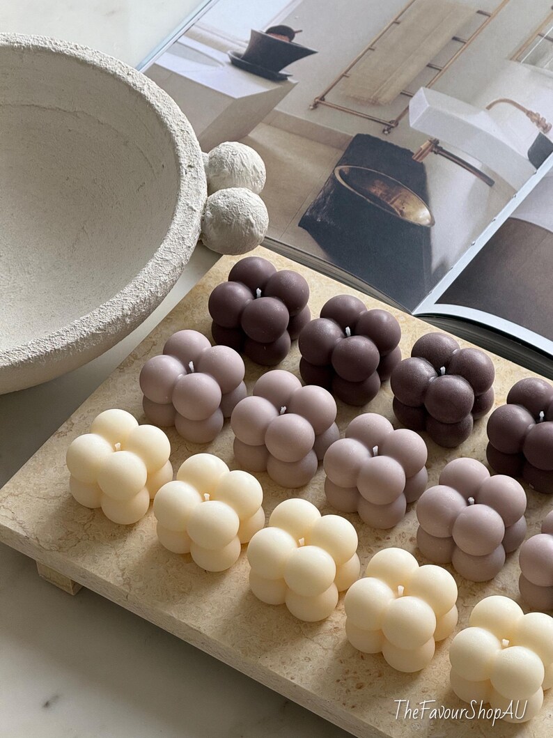 May include: A selection of bubble-shaped candles in cream, mauve, and brown hues. The candles are displayed on a light-toned rectangular tray. A textured bowl and decorative spheres are visible in the background.
