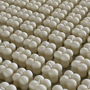 May include: A close-up of many ivory-coloured bubble candles. Each candle has a spherical shape with four rounded sections and a single wick. The candles are arranged in neat rows, creating a visually appealing pattern.