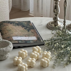 May include: Ivory-coloured bubble candles are displayed on a marble surface, alongside an open book with the text "Inspiring Your Journey". Silver candlesticks and sprigs of greenery are also present. The Favour Shop AU is visible in the bottom right corner.