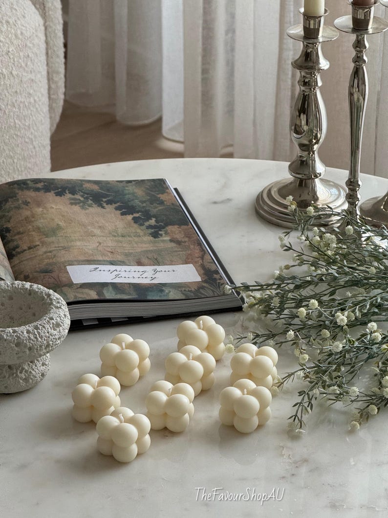 May include: Ivory-coloured bubble candles are displayed on a marble surface, alongside an open book with the text "Inspiring Your Journey". Silver candlesticks and sprigs of greenery are also present. The Favour Shop AU is visible in the bottom right corner.