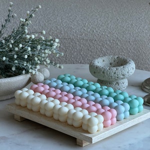 May include: A selection of pastel-coloured bubble candles, including white, pink, blue, and green, presented on a light wooden tray. The candles feature a distinctive, rounded design and are displayed on a marble surface.
