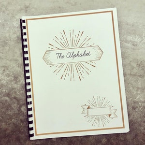 Op de afbeelding: Een wit notitieboek met een gouden rand en een gouden zonnestralenontwerp. Het notitieboek heeft een zwarte spiraalbinding en de tekst "The Alphabet" in een gouden banner.