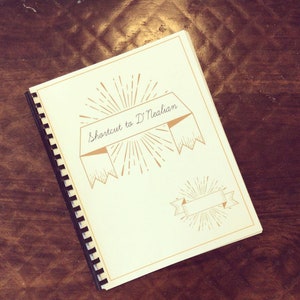 Peut inclure: Un carnet à spirales blanc avec le titre "Shortcut to D'Nealian" en écriture cursive. Le titre est entouré d'une bannière décorative et d'un motif de soleil.