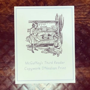 Op de afbeelding: Een zwart-witte illustratie van twee meisjes in een bibliotheekomgeving. De afbeelding heeft de titel "McGuffey's Third Reader Copywork: D'Nealian Print".