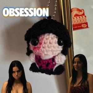 May include: A small, handmade crochet doll keychain featuring black yarn hair, large black plastic eyes, and a pale pink face with red accents. The doll wears a pink and black patterned outfit. The word OBSESSION is printed in white above the doll.
