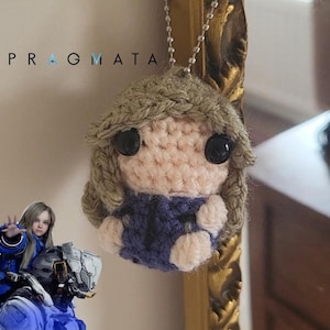 May include: A small crochet doll ornament featuring light brown yarn hair, a peach-colored face with black bead eyes, and a dark blue outfit. The figure hangs from a silver ball chain and is displayed near the text PRAGMATA and a small character image.