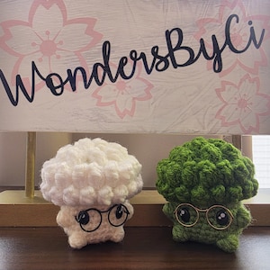Puede incluir: Dos figuras de verduras de ganchillo, una coliflor blanca y un brócoli verde, cada una con gafas redondas pequeñas. El fondo presenta un letrero con el texto "WondersByCi" y diseños de flores rosas.