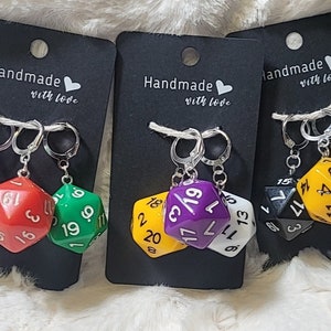 May include: Earrings featuring three multi-sided dice in various colors: purple, red, green, yellow, white, and black. Each earring set is attached to a black card with the text "Handmade with love."