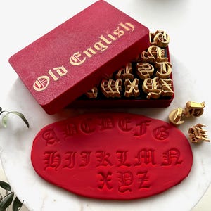 Könnte beinhalten: Eine rote Schachtel mit goldfarbenem Schriftzug "Old English" enthält Holzbuchstabenstempel. Die Stempel sind in verschiedenen altenglischen Schriftarten gehalten. Ein rotes Oval aus Ton zeigt gestempelte Buchstaben.