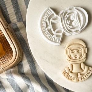 Puede incluir: Dos cortadores de galletas de plástico blanco con forma de astronauta con el texto "Future is Female" en la parte inferior. El astronauta lleva un casco y tiene una expresión decidida en su rostro.