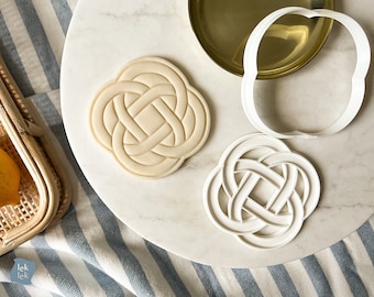 May include: Two white plastic cookie cutters in the shape of a Celtic knot. The cutters are on a white marble surface.