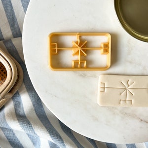 Puede incluir: Primer plano de un cortador de galletas y una galleta con un diseño geométrico. El cortador de galletas está hecho de plástico y es de color amarillo. La galleta está hecha de masa y es de color blanco.