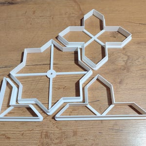 May include: A collection of white plastic cookie cutters in various geometric shapes, including stars and crosses, arranged on a wooden surface. The cutters are designed for baking and crafting.