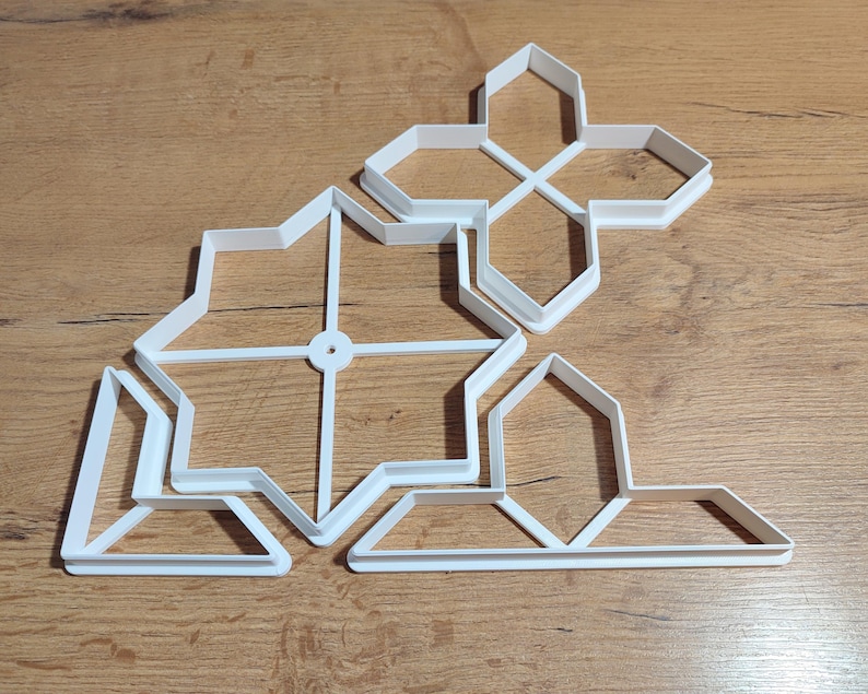 May include: A collection of white plastic cookie cutters in various geometric shapes, including stars and crosses, arranged on a wooden surface. The cutters are designed for baking and crafting.