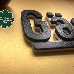 May include: Close-up of 3D-printed black letters with a textured surface, white accents, and a light brown base. The letters spell out a word. A green and white clover-shaped graphic with German text is in the upper left corner.