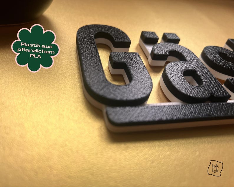 May include: Close-up of 3D-printed black letters with a textured surface, white accents, and a light brown base. The letters spell out a word. A green and white clover-shaped graphic with German text is in the upper left corner.