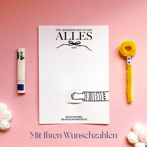 May include: A white rectangular card with black text reading F&Uuml;R JEMANDEN DER SCHON ALLES HAT and BAUCHNABEL REINIGUNGSB&Uuml;RSTE, featuring a graphic of a hand. Beside it is a rolled banknote and a yellow pipe cleaner with the number 40 on beads.