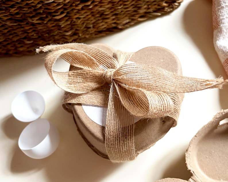 K&ouml;nnte beinhalten: Eine herzf&ouml;rmige, hellbraune Geschenkbox, die mit einem nat&uuml;rlichen Jutegarnband und einer Schleife gebunden ist. Zwei wei&szlig;e, eierschalenf&ouml;rmige Beh&auml;lter befinden sich neben der Box. Die Box steht auf einer wei&szlig;en Oberfl&auml;che.