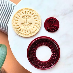 Puede incluir: Tres cortadores de galletas, dos son rojos y uno es beige. El cortador beige es un círculo con un diseño de martillo y un patrón en zigzag. Los cortadores rojos son un círculo con un patrón en zigzag similar y un diseño de martillo más pequeño.