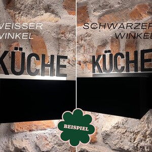 May include: A black and white kitchen sign with the word "K&Uuml;CHE" in large letters. The sign is mounted on a black rectangular base, with the text "WEISSER WINKEL" above the word "K&Uuml;CHE". The background is a brick wall.