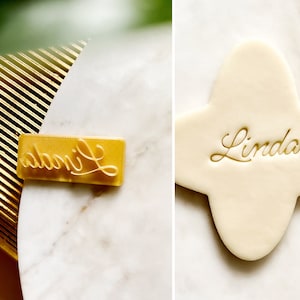 May include: A gold-coloured plastic stamp with the name "Linda" embossed on it. The stamp is on a white marble surface.