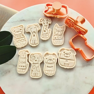 Puede incluir: Un conjunto de cortadores de galletas con forma de perro y galletas horneadas. Los cortadores son de color cobre y las galletas son de color beige. Las galletas tienen varios dise&ntilde;os de perros, incluyendo un perro con su&eacute;ter.