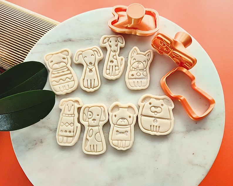 Puede incluir: Un conjunto de cortadores de galletas con forma de perro y galletas horneadas. Los cortadores son de color cobre y las galletas son de color beige. Las galletas tienen varios dise&ntilde;os de perros, incluyendo un perro con su&eacute;ter.