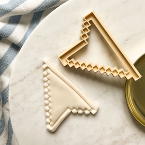 May include: A beige plastic cookie cutter in the shape of a pennant flag, next to a cookie with the same shape. The cookie cutter has a geometric design along the edges. The cookie is light beige and has a textured surface.
