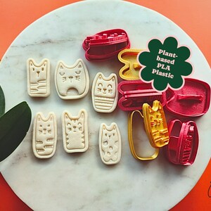 May include: A collection of cat-shaped cookie cutters and stamped cookies. The cutters are red and yellow, while the cookies are beige. The image includes a green flower-shaped graphic that reads "Plant-based PLA Plastic".