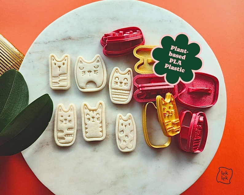 May include: A collection of cat-shaped cookie cutters and stamped cookies. The cutters are red and yellow, while the cookies are beige. The image includes a green flower-shaped graphic that reads "Plant-based PLA Plastic".