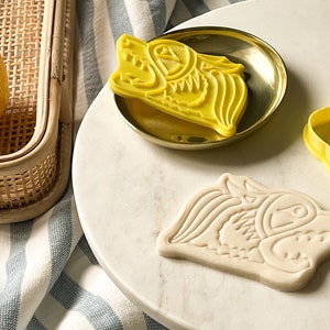 Puede incluir: Dos cortadores de galletas de plástico amarillo en forma de cabeza de lobo. Un cortador está en un plato dorado y el otro en una superficie blanca. Una galleta blanca con el diseño de la cabeza de lobo está en la superficie blanca.