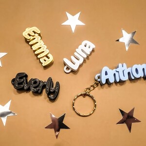 May include: Five personalized keychains with names spelled out in 3D letters. The names are "Emma", "Luna", "Anthony", "Every", and a fifth keychain with a gold ring.