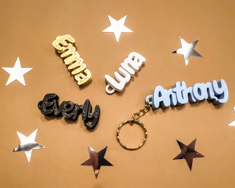 May include: Five personalized keychains with names spelled out in 3D letters. The names are "Emma", "Luna", "Anthony", "Every", and a fifth keychain with a gold ring.