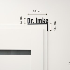 May include: Black metal sign that reads "Dr. Imke" with dimensions of 28 cm wide and 8.5 cm high. The sign is mounted on a corner piece that is 11.5 cm long. The sign is displayed on a white wall.