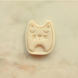 May include: A light beige cat-shaped cookie cutter with a smiling face. The cutter has a rectangular shape with rounded edges and a small opening at the bottom. The cat's eyes and nose are indented.