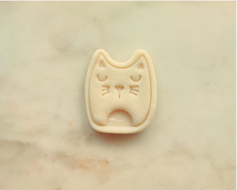 May include: A light beige cat-shaped cookie cutter with a smiling face. The cutter has a rectangular shape with rounded edges and a small opening at the bottom. The cat's eyes and nose are indented.