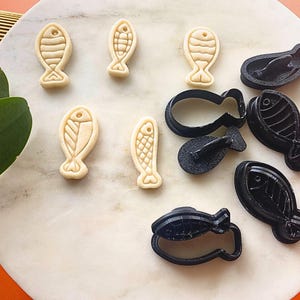 Könnte beinhalten: Draufsicht auf Fisch-Ausstecher und Kekse. Die Kekse sind hellbeige und weisen detaillierte Fischmuster auf. Schwarze Kunststoffausstecher in verschiedenen Fischformen sind ebenfalls vorhanden, zusammen mit den Keksen. Die Artikel sind auf einer weißen Marmoroberfläche angeordnet.