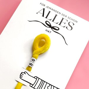 Op de afbeelding: Een witte wenskaart met de zwarte tekst F&Uuml;R JEMANDEN DER SCHON ALLES HAT en een afbeelding van een hand die een gele, pluizige pijpenrager vasthoudt. De pijpenrager is aan de bovenkant gelust en bevat twee witte vierkante kralen met het getal 50.