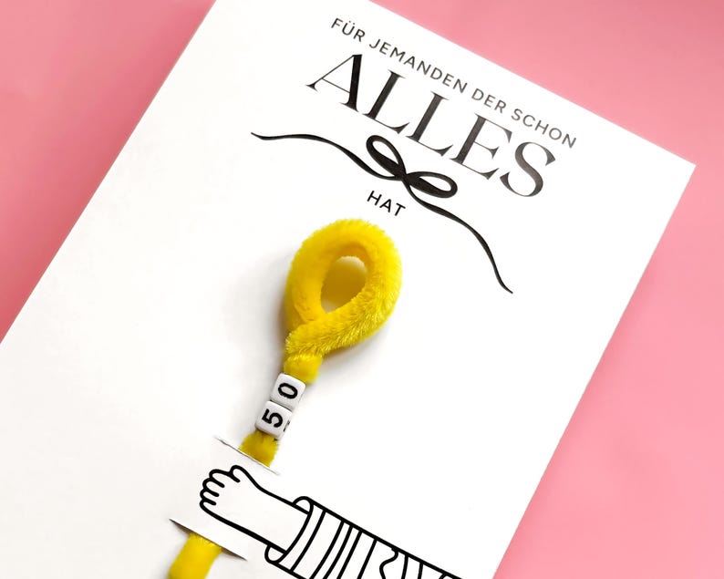 Op de afbeelding: Een witte wenskaart met de zwarte tekst F&Uuml;R JEMANDEN DER SCHON ALLES HAT en een afbeelding van een hand die een gele, pluizige pijpenrager vasthoudt. De pijpenrager is aan de bovenkant gelust en bevat twee witte vierkante kralen met het getal 50.