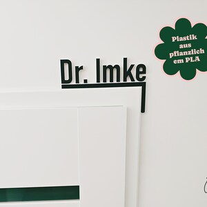 May include: Black sign with the text "Dr. Imke" on a white wall. A green flower-shaped sticker with the text "Plastik aus pflanzlich em PLA" is next to the sign. The sign is mounted on a white door.