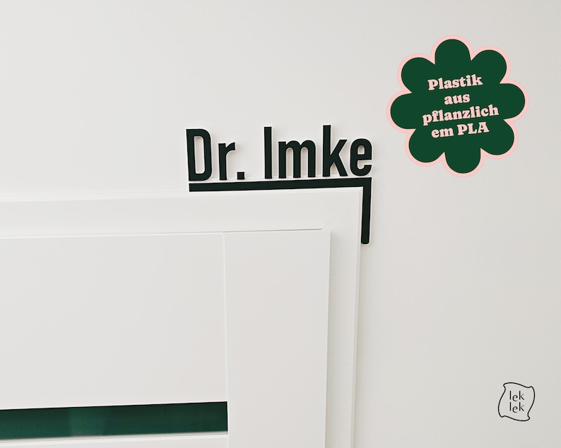 May include: Black sign with the text "Dr. Imke" on a white wall. A green flower-shaped sticker with the text "Plastik aus pflanzlich em PLA" is next to the sign. The sign is mounted on a white door.