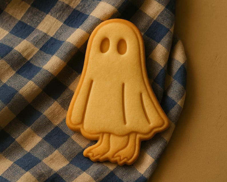 May include: A Halloween-themed ghost-shaped cookie with a light brown color. The cookie has two oval eye indentations and a smiling mouth. It rests on a blue and white checkered cloth.