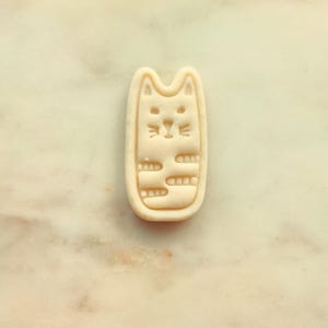 May include: A beige cat-shaped cookie cutter. The cutter has a rectangular shape with rounded edges and a cat face design. The cat's face includes eyes, a nose, and whiskers. The body has horizontal lines.