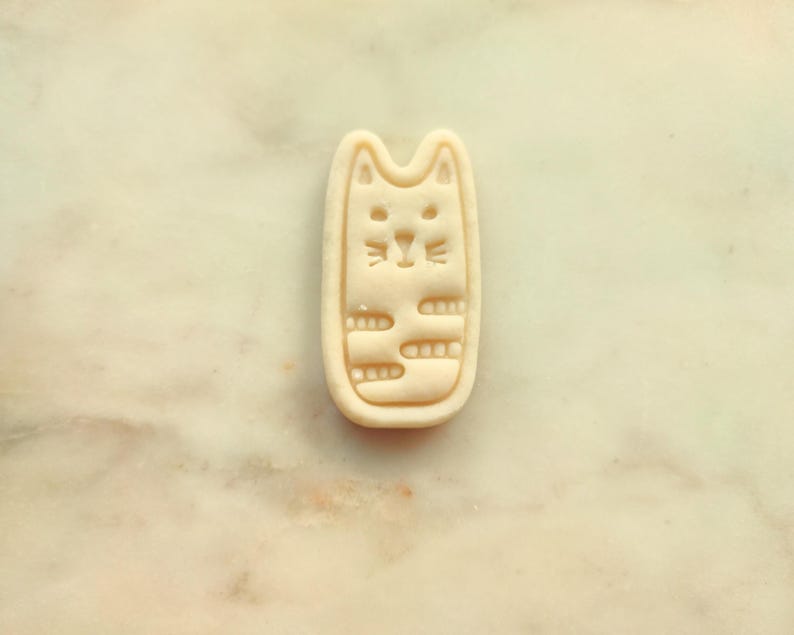 May include: A beige cat-shaped cookie cutter. The cutter has a rectangular shape with rounded edges and a cat face design. The cat's face includes eyes, a nose, and whiskers. The body has horizontal lines.