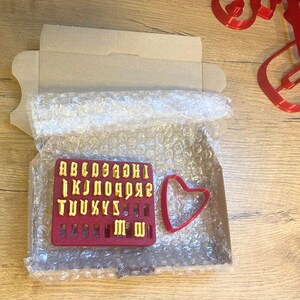 May include: A red rectangular plastic mold containing yellow raised letters of the alphabet from A to Z, accompanied by a separate red heart-shaped cookie cutter, all packaged inside a cardboard box lined with clear bubble wrap.
