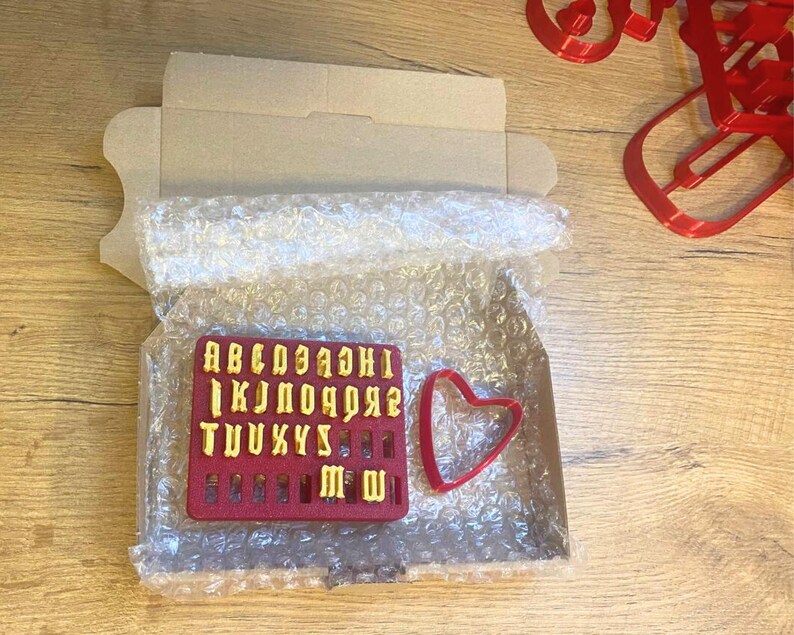 May include: A red rectangular plastic mold containing yellow raised letters of the alphabet from A to Z, accompanied by a separate red heart-shaped cookie cutter, all packaged inside a cardboard box lined with clear bubble wrap.