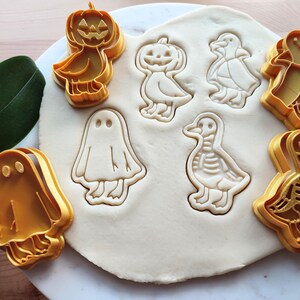 May include: A collection of orange plastic cookie cutters in Halloween and autumn shapes, including a pumpkin, ghost, and ducks. The cutters are pressed into a sheet of dough, creating impressions of the designs.