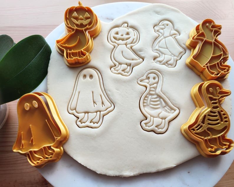 May include: A collection of orange plastic cookie cutters in Halloween and autumn shapes, including a pumpkin, ghost, and ducks. The cutters are pressed into a sheet of dough, creating impressions of the designs.