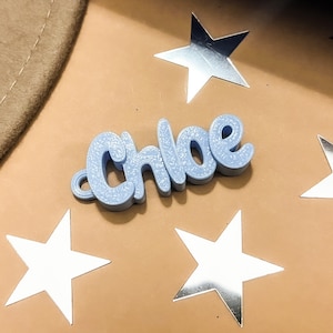 May include: A light blue, 3D printed name tag with the name "Chloe" spelled out in a cursive font. The name tag is sitting on a brown surface with silver stars scattered around it.