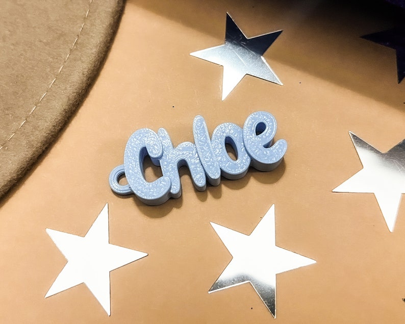 May include: A light blue, 3D printed name tag with the name "Chloe" spelled out in a cursive font. The name tag is sitting on a brown surface with silver stars scattered around it.